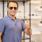 Indian man wear sunglasses and show thumbs up with background of eyeglasses shelves